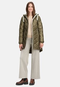 Olijfgroene gewatteerde jas met een capuchon, voorzien van ritszakken en een middellange snit. Gecombineerd met geruite wijde broek en bruine enkellaarzen.