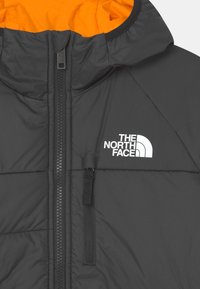 Musta eristyspuku, jossa on kirkkaan oranssi sisus, täysipitkä vetoketjusulku ja logovaihtoehto rinnassa. Rakenteeltaan kohokuvioitu, pehmustettu kangas.