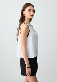 Haut sans manches en lin blanc avec des détails noirs, associé à un short noir. Le look comprend une coupe décontractée et une texture subtile. Un bracelet doré est visible.