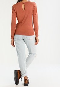 Haut à manches longues couleur rouille avec une ouverture en trou de serrure dans le dos, associé à un jean bleu clair aux revers retroussés et des chaussures à plateforme marron.