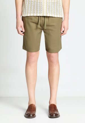 Persona che indossa pantaloncini verde oliva con coulisse, una maglietta bianca con motivo e scarpe slip-on in pelle marrone, in piedi su un pavimento bianco.