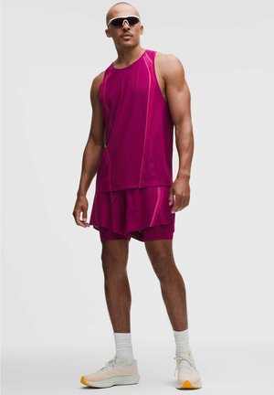 Muskelbepackter Mann trägt ein dunkelrosa ärmelloses Oberteil und passende Shorts, weiße Socken, beige Sneakers und schwarze Sportsonnenbrille, er steht.