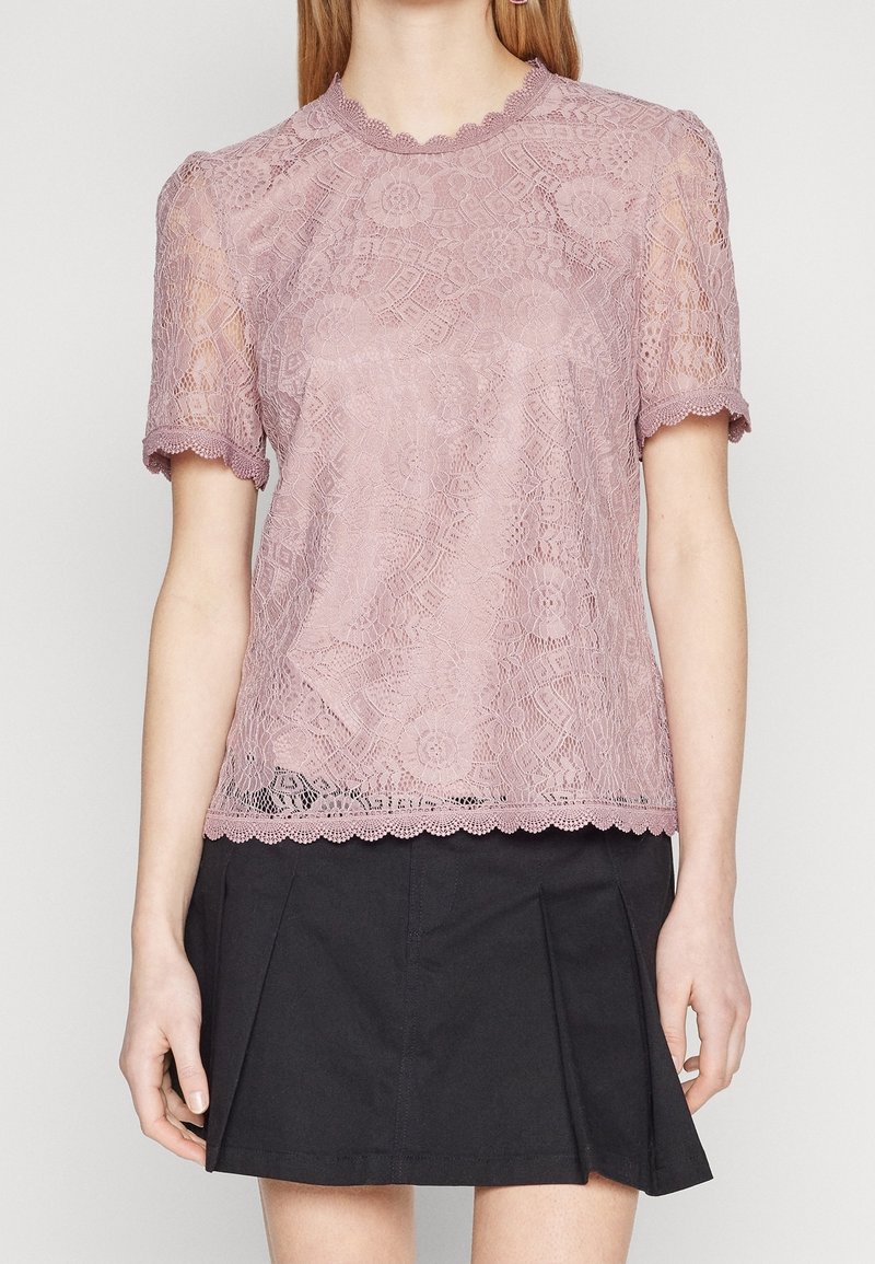 Femme portant un haut en dentelle mauve à manches courtes avec des motifs floraux et des bords festonnés, associé à une mini-jupe plissée noire.
