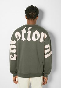 Olivgrüner Sweatshirt mit Oversized-Schnitt, versehen mit großen rosa Buchstaben auf dem Rücken und den Ärmeln. Weicher Stoff mit gerippten Bündchen und Saum.
