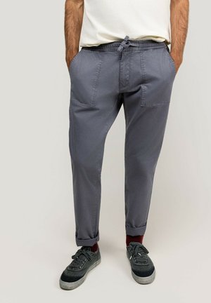 Hombre con pantalones casuales grises con puños enrollados, zapatillas oscuras y camisa blanca, de pie con las manos en los bolsillos.