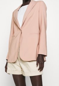 Blazer rosa empolvado con solapa de muesca, un solo botón y bolsillos delanteros. Llevado sobre una blusa blanca y combinado con pantalones cortos crema. Tela suave.
