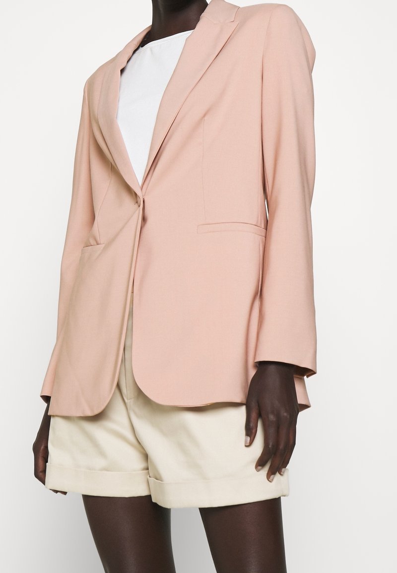 Blazer rosa empolvado con solapa de muesca, un solo botón y bolsillos delanteros. Llevado sobre una blusa blanca y combinado con pantalones cortos crema. Tela suave.