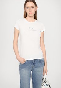 T-shirt bianca in cotone con maniche corte, caratterizzata da un logo stampato grigio chiaro "A|X ARMANI EXCHANGE" al centro. Abbinata a jeans in denim blu.