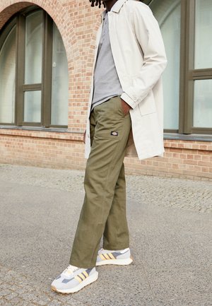 Cappotto leggero beige, polo grigia e pantaloni verde oliva. Sneakers grigie e gialle con suola texturizzata. Ambiente urbano con sfondo di mattoni.