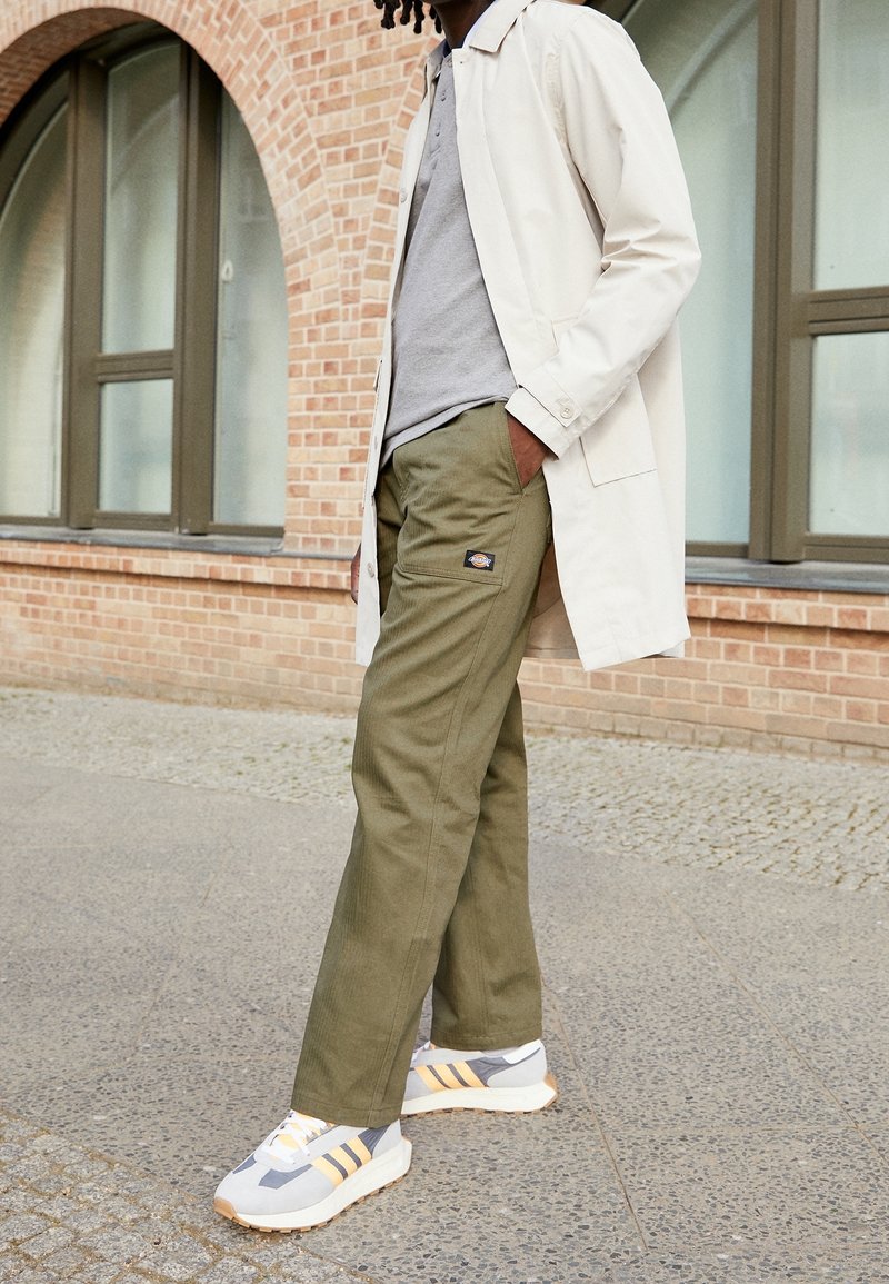 Cappotto leggero beige, polo grigia e pantaloni verde oliva. Sneakers grigie e gialle con suola texturizzata. Ambiente urbano con sfondo di mattoni.