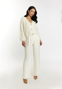 Cardigan morbido e soffice con dettagli di bottoni, abbinato a pantaloni a gamba larga bianchi crema e décolleté nudi a punta. Texture liscia.
