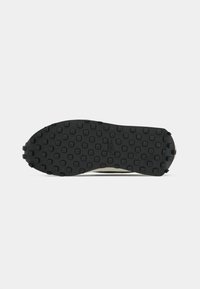 Semelle extérieure en caoutchouc noir avec des motifs carrés et circulaires, texturée pour l'adhérence. Tige en tissu clair avec un design minimaliste et sans marquage visible.