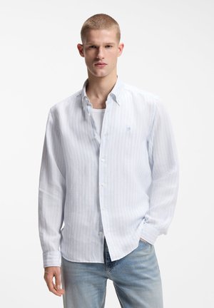 Νεαρός άνδρας που φοράει ένα γαλάζιο ριγέ πουκάμισο με κουμπιά πάνω από ένα λευκό μπλουζάκι και γαλάζια τζιν, στέκεται με το ένα χέρι στην τσέπη.