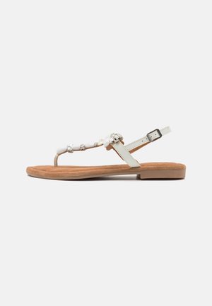 Flad tan sandal med hvid klip-strap pyntet med rhinsten og en justerbar hvid ankelrem med spænde.