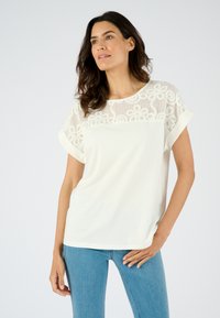 Top blanco de manga corta con un borde de encaje y patrones florales en la parte del pecho, tejido suave y un ajuste relajado. Combinado con jeans azules.