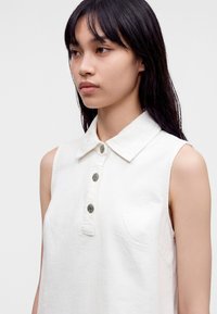 Jeune femme aux longs cheveux foncés portant une chemise blanche sans manches avec col et trois boutons métalliques, regardant vers la gauche.