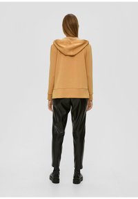 Haut à capuche marron clair avec capuche et poignets côtelés, associé à un pantalon en simili cuir noir. Design simple, texture lisse et silhouette ajustée.