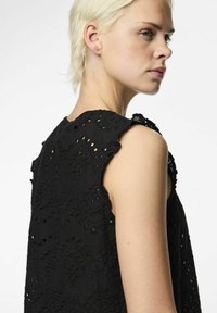 Top sans manches noir avec une broderie complexe, des épaules à bords festonnés et un design de tissu texturé avec des détails découpés.