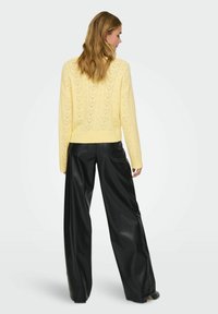 Pull jaune en tricot avec un motif en V et un ourlet côtelé, associé à un pantalon large en similicuir noir. Détails texturés visibles.