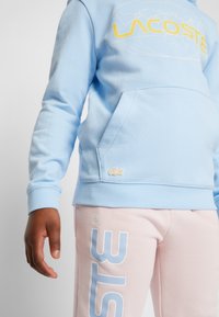 Ljusblå sweatshirt med en stor gul Lacoste-logotyp och världskarta. Har en framficka och broderad logotyp på kanten.