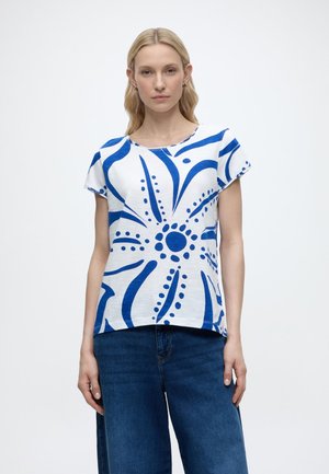 Blonde vrouw die een wit shirt met korte mouwen draagt met een gedurfde blauwe abstracte bloemenprint en brede blauwe jeans, staand tegen een witte achtergrond.