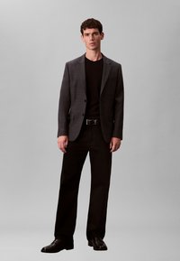 Blazer gris texturé sur une chemise noire, assorti à un pantalon droit noir et des chaussures noires. Le modèle pose devant un fond uni.