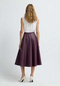Kvinde med krøllet blond hår iført en hvid ærmeløs top, lilla midi læderskirt og hvide højhælede sko, stående foran en ensfarvet baggrund.