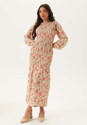 Kvinde med langt mørkt hår iført en cremet, langærmet blomsterkjole og beige hæle, som står med hænderne på hofterne foran en hvid baggrund.