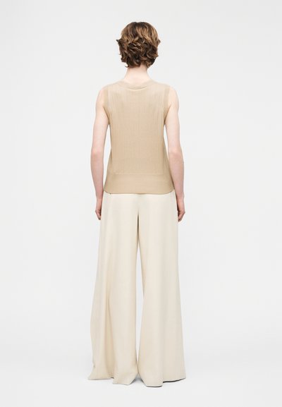 Άτομο με κοντά κυματιστά μαλλιά, φοράει μπεζ τοπ χωρίς μανίκια και εκρού παντελόνι φαρδύ, στέκεται με την πλάτη προς το μέρος μας.