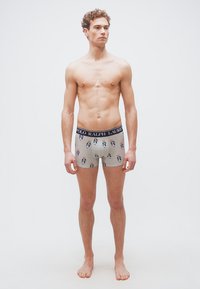 Νεαρός άνδρας που στέκεται ξυπόλυτος, φορώντας γκρι εσώρουχα boxer με μικρά σχέδια αρκούδας και μαύρη ζώνη Polo Ralph Lauren.