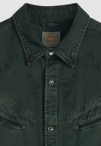 Camisa de mezclilla verde oscuro con cuello puntiagudo, detalles bordados en los hombros, cierre frontal de botones y acentos decorativos de costura negra.