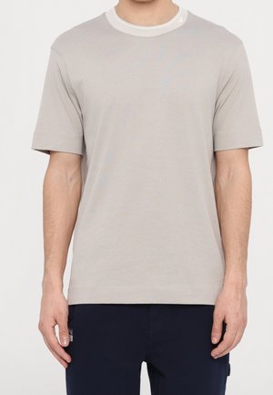 Mann trägt ein hellbeiges T-Shirt mit kurzen Ärmeln und Rundhalsausschnitt sowie dunkelblaue Hosen vor weißem Hintergrund.