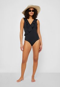 Maillot de bain noir une pièce avec un design à col en V froncé, présentant une silhouette ajustée et une texture lisse. Porté avec un chapeau de paille à large bord et des lunettes de soleil.