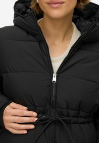 Ženska ruka podešava vrpcu na crnoj pufer jakni s kapuljačom, djelomično zatvorenoj preko svijetle košulje.