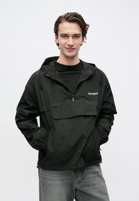 Νεαρός άνδρας που φοράει μαύρο αδιάβροχο με κουκούλα, εμπρόσθιο φερμουάρ και τσέπη, στέκεται με τα χέρια στις τσέπες μπροστά σε μια απλή φόντο.