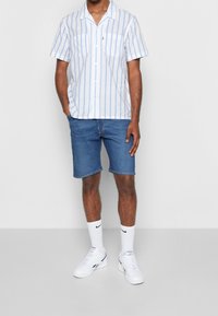 Camisa de manga corta a rayas blancas y azul claro con botones, combinada con pantalones cortos de mezclilla azul oscuro. Zapatillas blancas con detalles en negro.