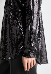 Black sequin top featuring a textured surface, long sleeves, and rounded hem. The fabric has a glossy finish with shimmering sequins.