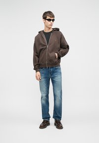 Sudadera marrón con cremallera y bolsillo delantero, camiseta negra debajo, vaqueros de lavado claro y zapatos marrón oscuro. Modelo con gafas de sol.