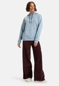 Sudadera de color azul claro con un bolsillo delantero, capucha con cordón y detalle del logo en verde. Combinada con pantalones anchos de pana oscura y zapatillas blancas.