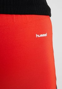 Röda sportleggings med en slät textur, som har en vit "hummel"-logotyp på sidan och ett svart ribbat midjeband.