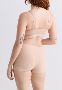 Frau mit heller Haut und roten Haaren, die einen beigen BH und Shorts trägt, zeigt Tattoos an ihrem linken und rechten Arm und steht vor einem einfarbigen hellen Hintergrund.