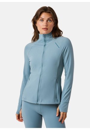 Donna con lunghi capelli castani che indossa una giacca con zip azzurra chiara e leggings abbinati, in piedi davanti a uno sfondo bianco.