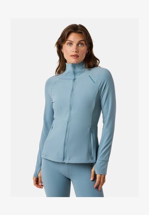 Vrouw met lang bruin haar, gekleed in een lichtblauw jack met ritssluiting en bijpassende leggings, staand tegen een witte achtergrond.