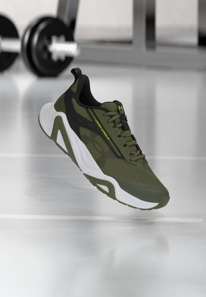 Зелена и черна спортна обувка Under Armour с бяло ходило, плаваща над отразяващия под на фитнес залата и размазани дъмбели на заден план.
