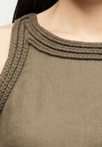 Κοντινό πλάνο ενός ατόμου που φοράει αμάνικο ελιάς κορυφή με πλεκτό διακόσμηση κατά μήκος του λαιμού και του ώμου.