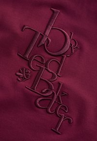 Tessuto rosso scuro in rilievo con la parola ricamata "HydrogradeBreaker" in grandi lettere sovrapposte e un piccolo simbolo floreale sotto.