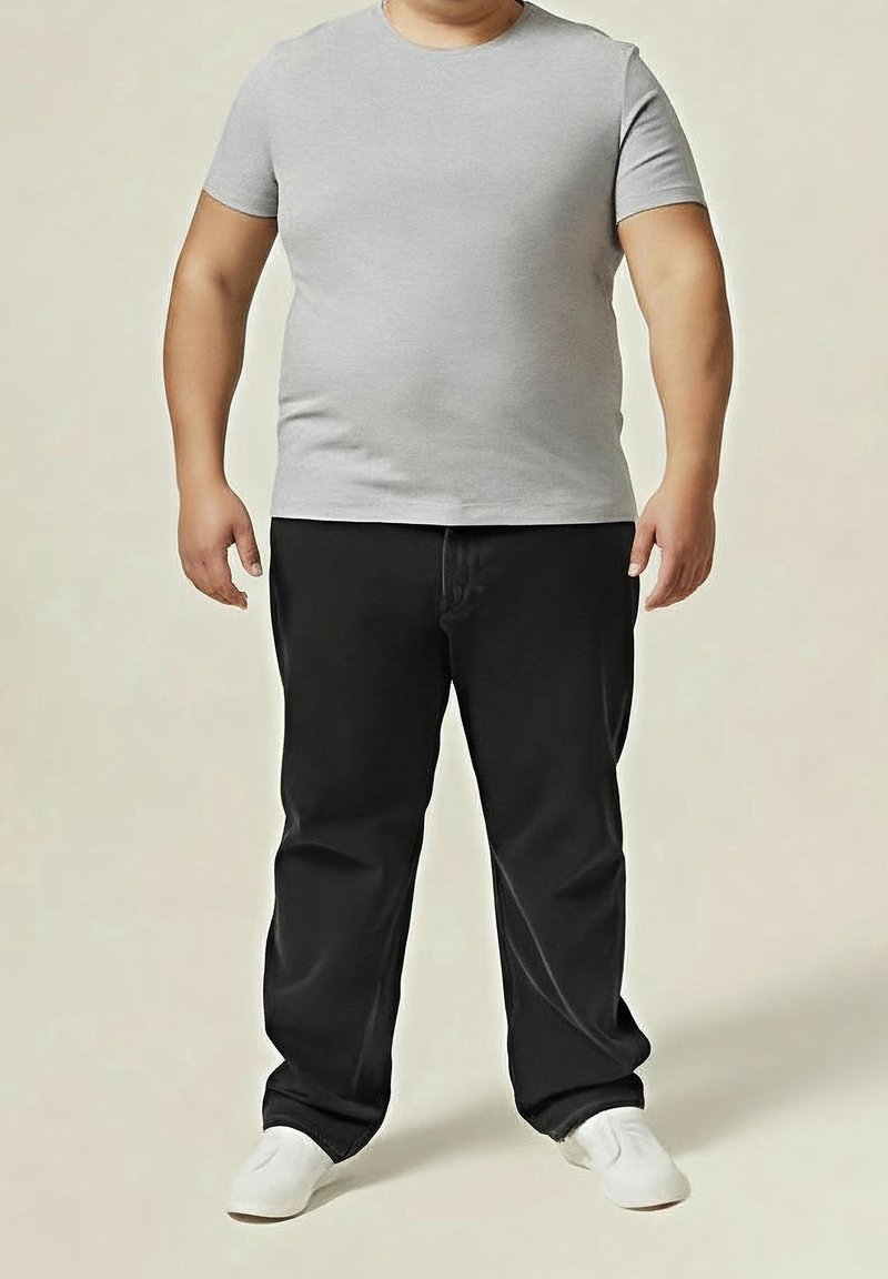 Adulte portant une chemise à manches courtes gris clair, un pantalon noir et des baskets blanches, debout devant un fond beige uni.