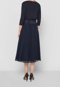 Woman wearing a navy lace-waist midi dress with a cropped three-quarter sleeve top and black high-heeled shoes, facing away.