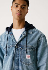 Spijkerjack met een marineblauwe hoodie eronder, lichtblauwe kleur, knoopsluiting aan de voorkant, borstzak en een Red Bull Racing logo patch.