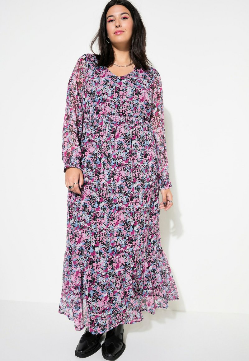 Robe longue et transparente à motifs floraux présentant des couleurs rose, bleu et noir, avec un décolleté en V, des manches longues et un ourlet volanté. Associée à des bottes noires.